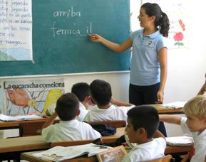 Students at a desk as teacher writes on a chalk board.