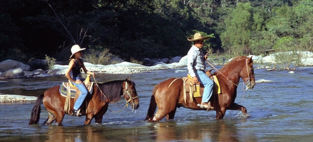 Two people on horseback crossing a river in Riviera Nayarit MX